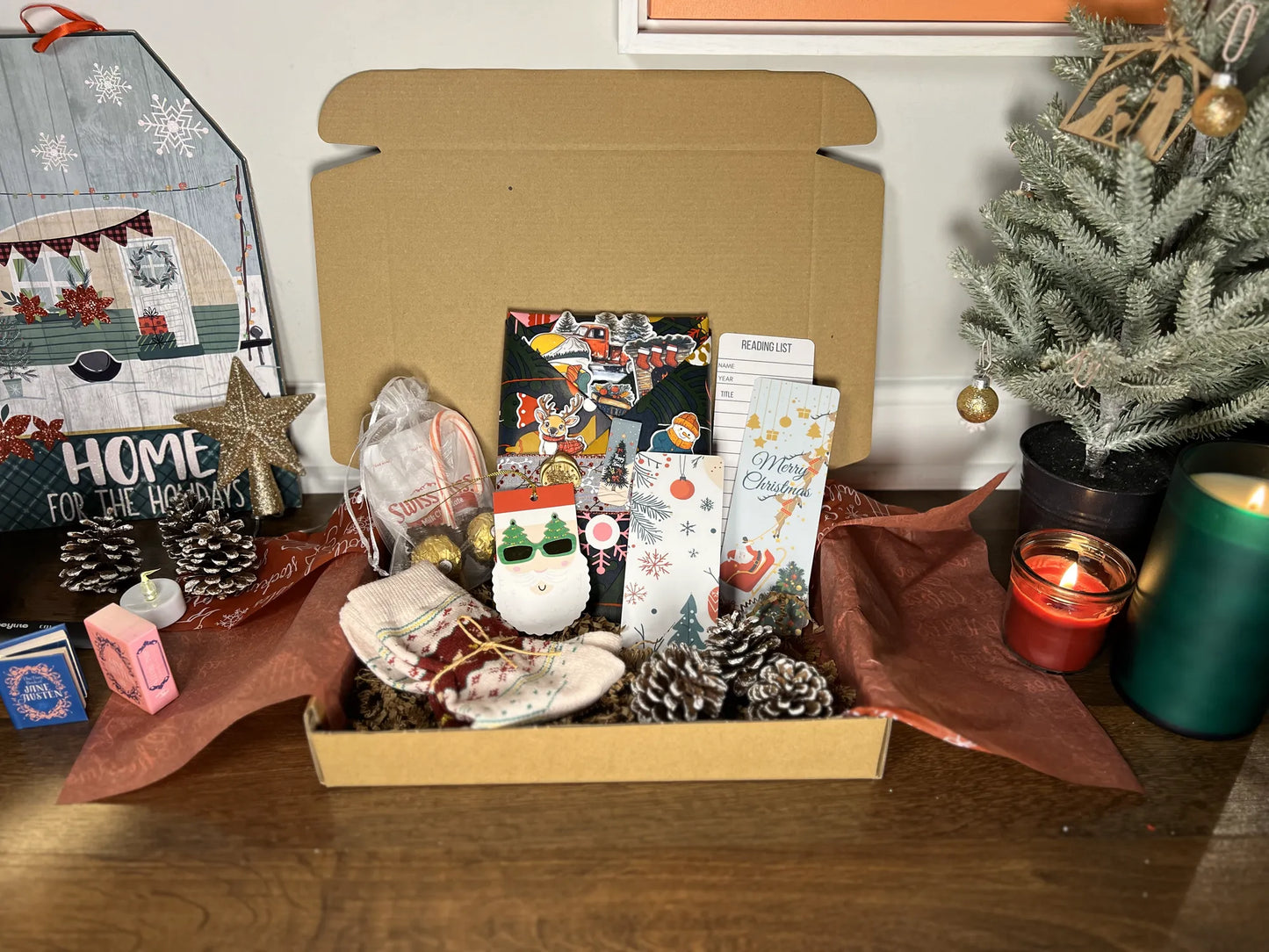 Christmas Blind Date with a Book Box with decorative items including a small tree, candles, and gift bags on a wooden surface.