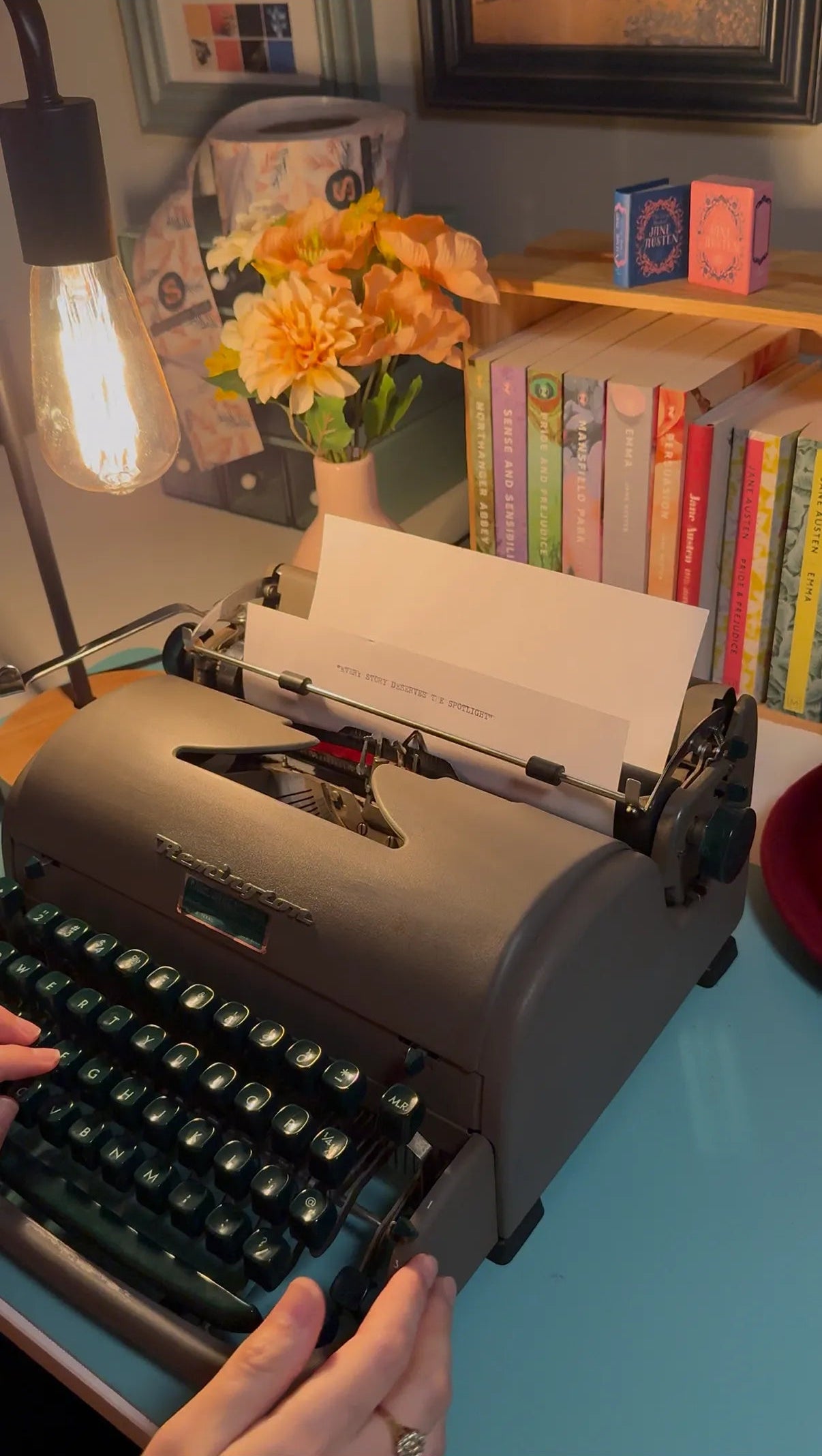 Person using a typewriter writes "Every Story Deserves the Spotlight" with a cozy setting including books and flowers.