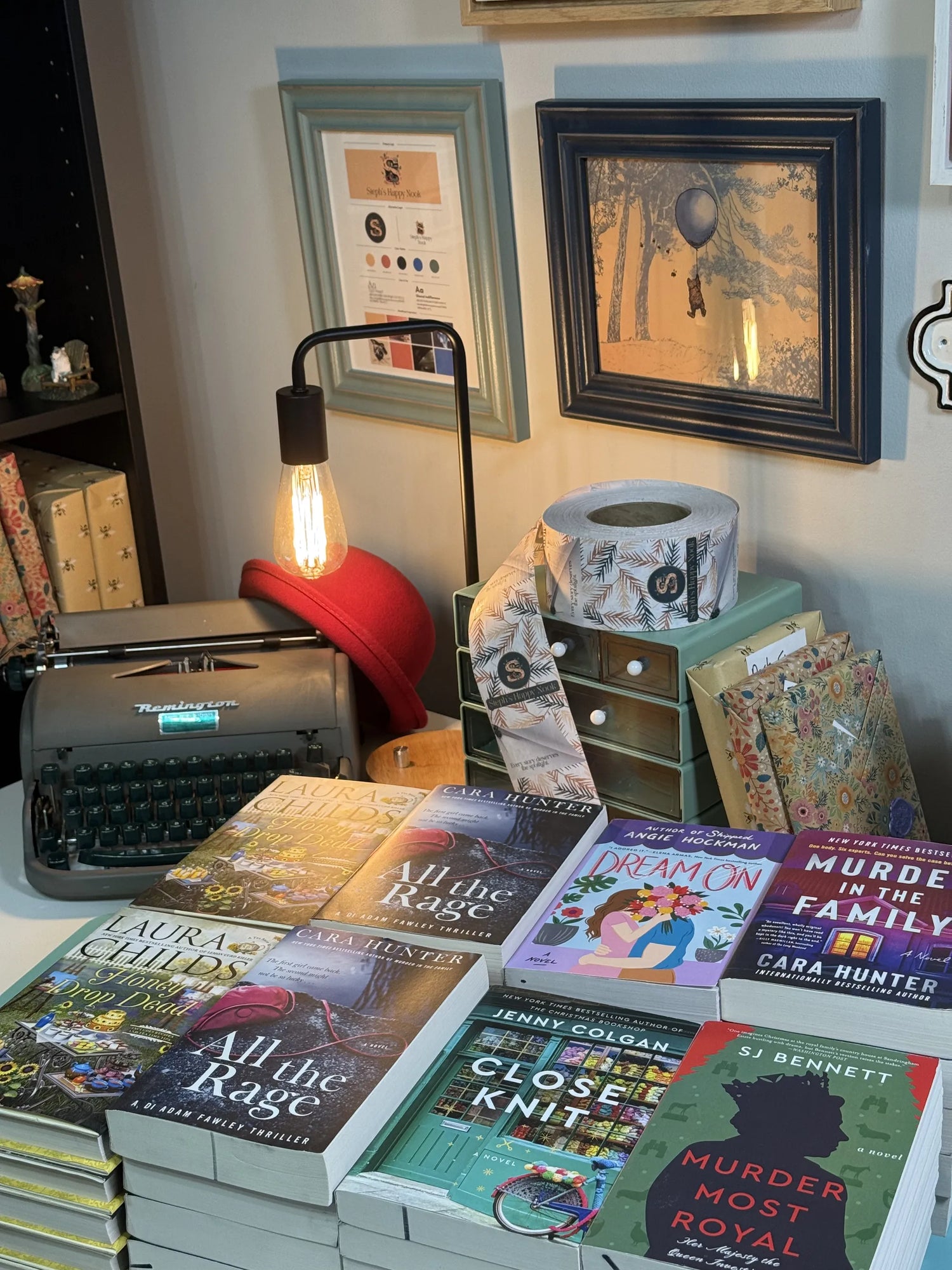 Stacks of books on a table with a typewriter and framed pictures in the background.