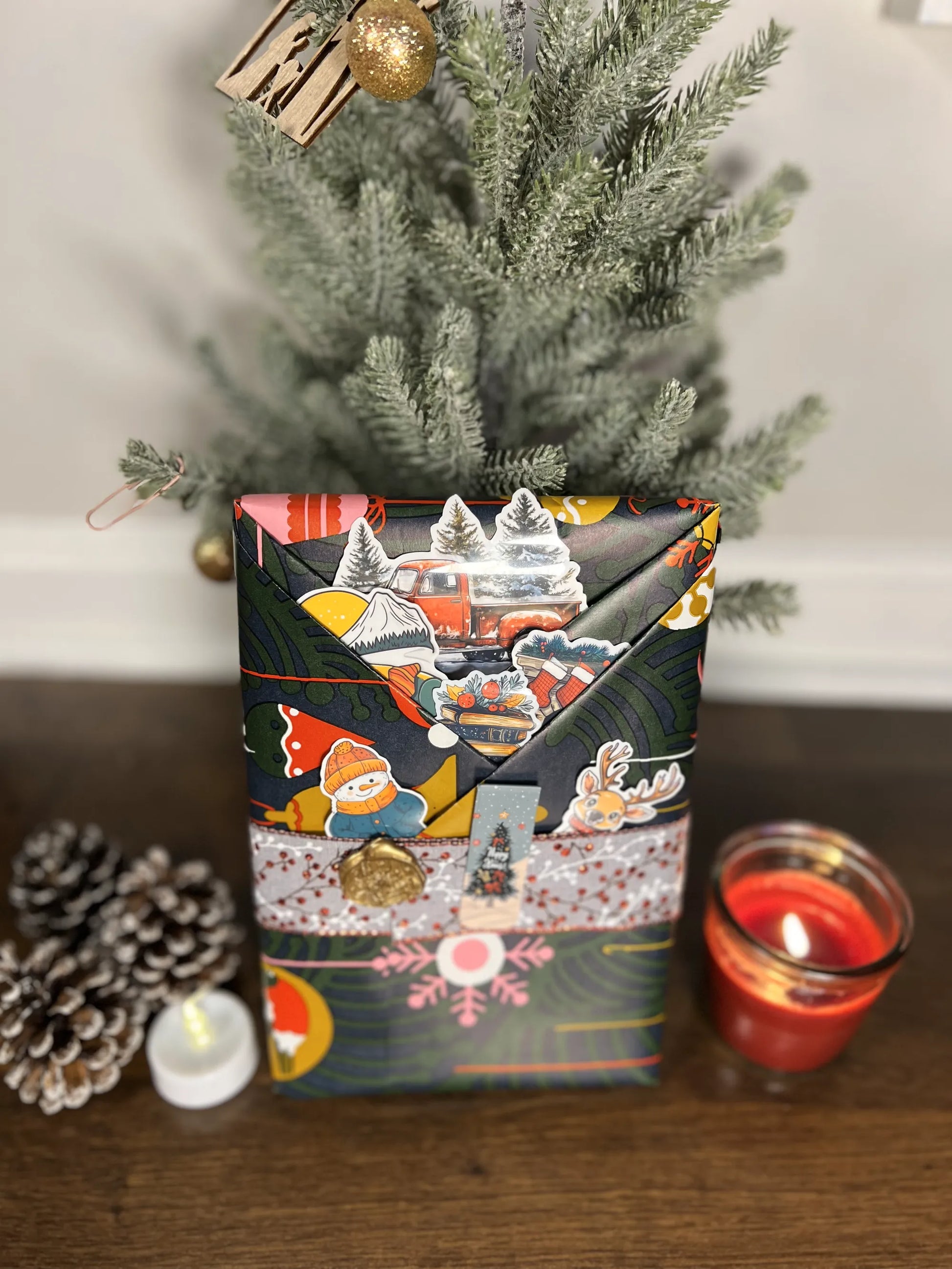 A wrapped mystery book with festive design next to a small Christmas tree and candles on a wooden surface.